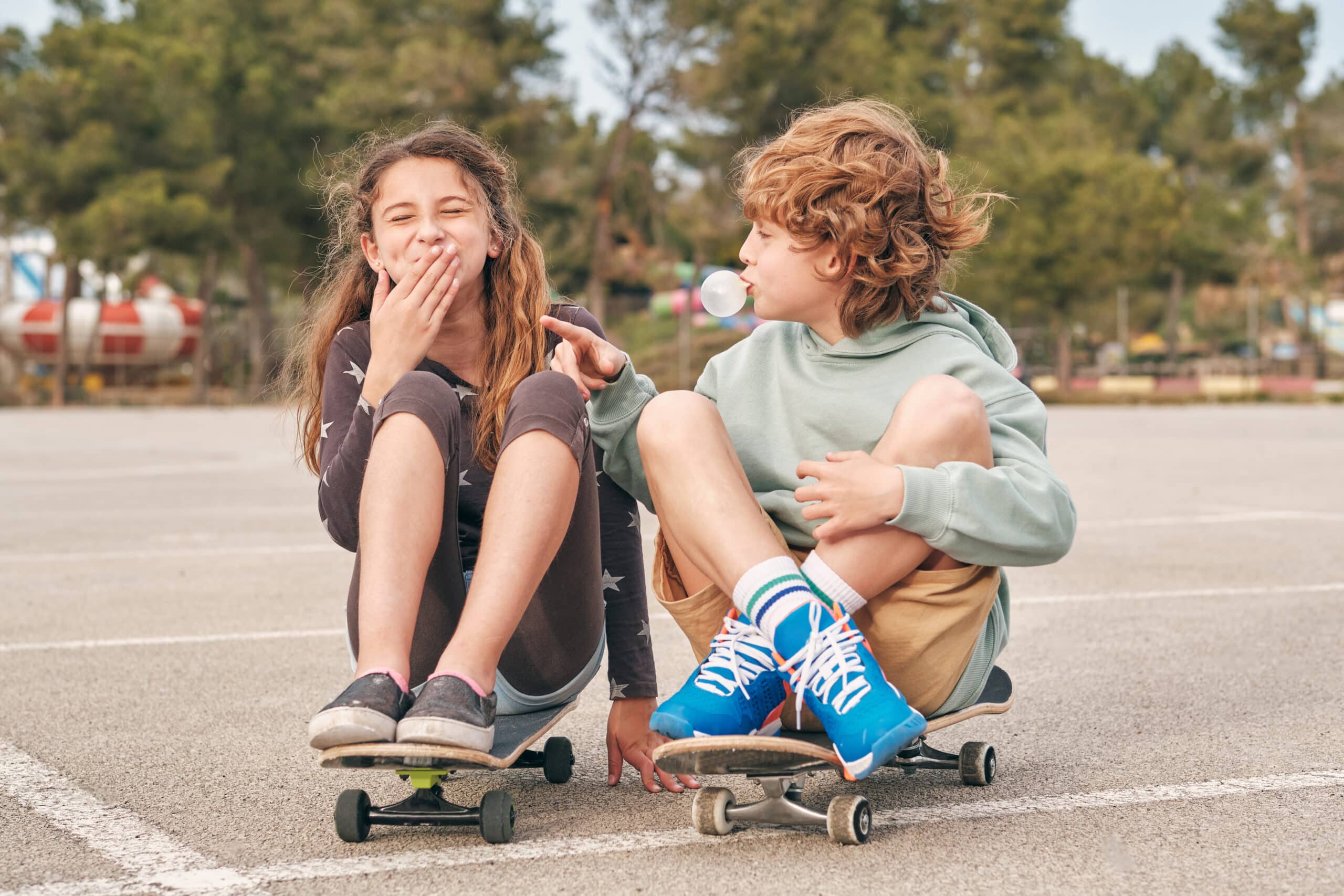 Delighted teenage boy and girl blowing chewing gum while sitting on skateboards in city and having fun together at weekend